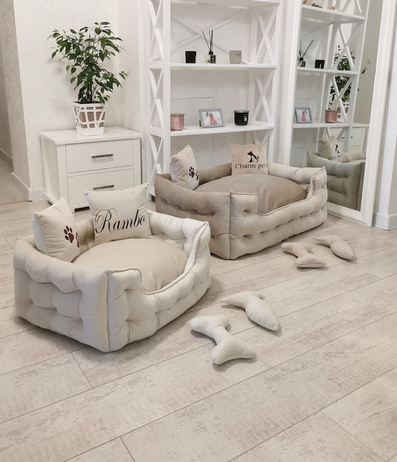 May include: Two beige dog beds with matching pillows and bone-shaped toys. One bed has the name "Rambo" embroidered on a pillow, and the other has "Charm pet" on a pillow. The beds have a tufted design.