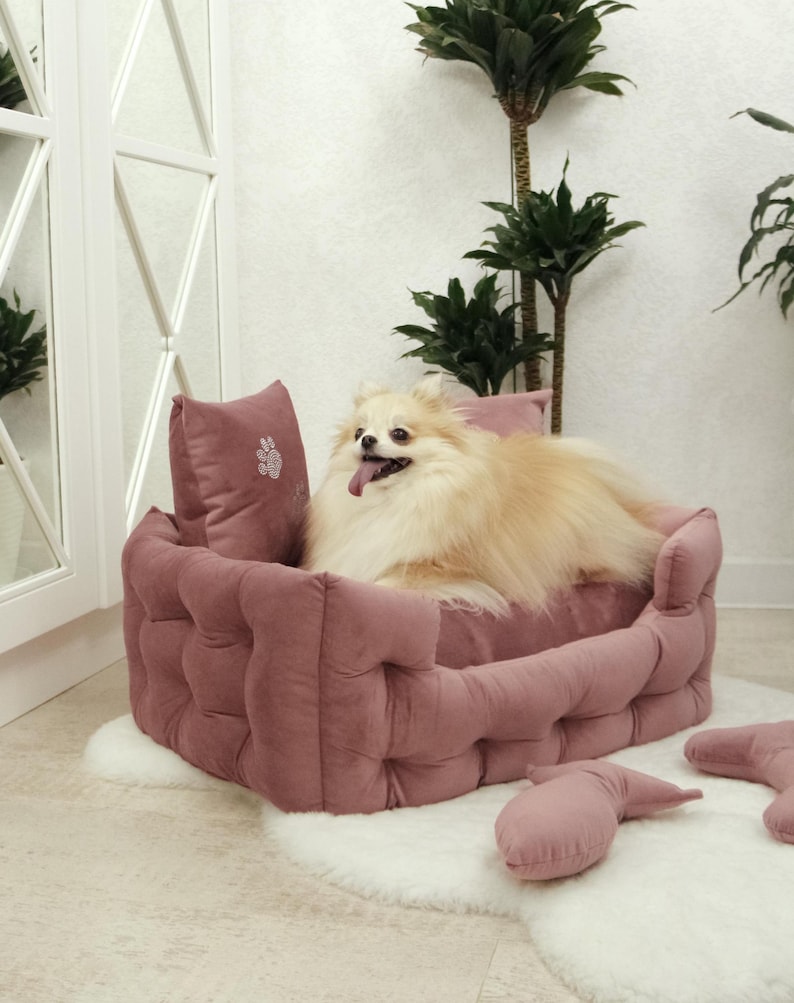 May include: A small, fluffy, light-colored dog rests in a dusty rose-colored, tufted pet bed. The bed sits on a white, fluffy rug. A matching pillow with a paw print is visible. Additional matching pillows are scattered around the bed.