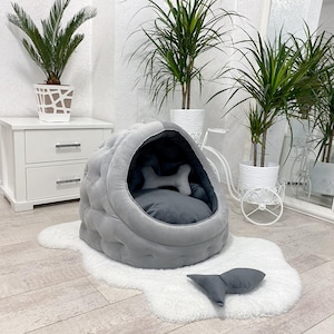 May include: A gray and white pet bed shaped like a dome with a plush interior and a matching gray pillow. The bed is on a white fluffy rug in a room with plants.