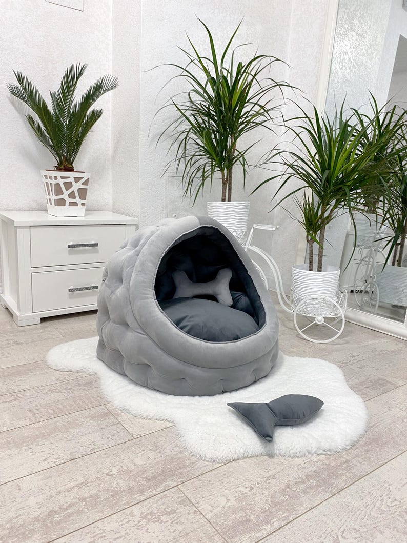 May include: A gray and white pet bed shaped like a dome with a plush interior and a matching gray pillow. The bed is on a white fluffy rug in a room with plants.