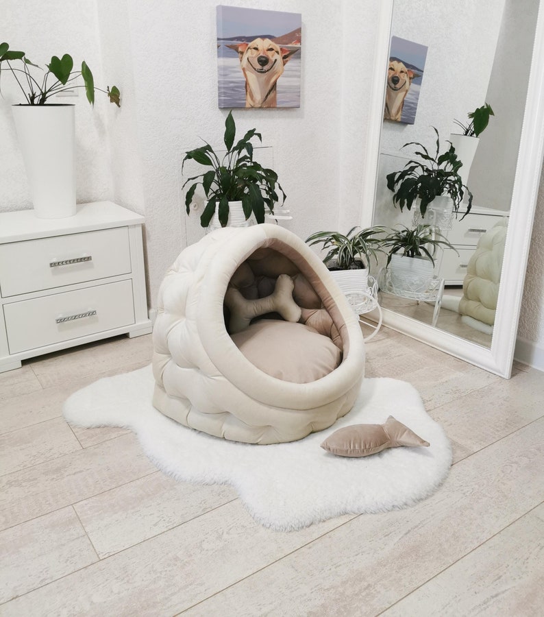 May include: A white and beige dog bed shaped like a dome with a plush, quilted exterior. The bed has a removable cushion and a bone-shaped toy inside. The bed is sitting on a white fluffy rug.