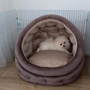 May include: A small white dog with fluffy fur is lying in a brown, round pet bed with a tufted design. The bed is in a room with a metal gate in the background.