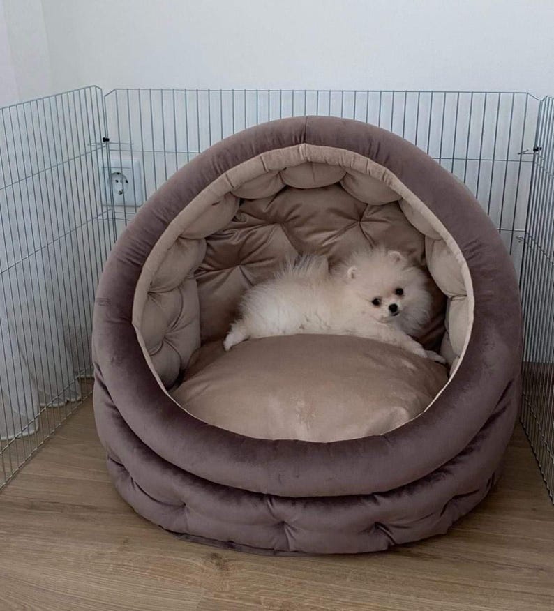 May include: A small white dog with fluffy fur is lying in a brown, round pet bed with a tufted design. The bed is in a room with a metal gate in the background.