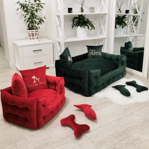 May include: Two pet beds, one red and one dark green, with matching pillows. The red bed has a pillow with the text "Charm Pets" and a dog silhouette. The green bed has a pillow with the name "Kieran". Several red and green bone-shaped toys are scattered around.