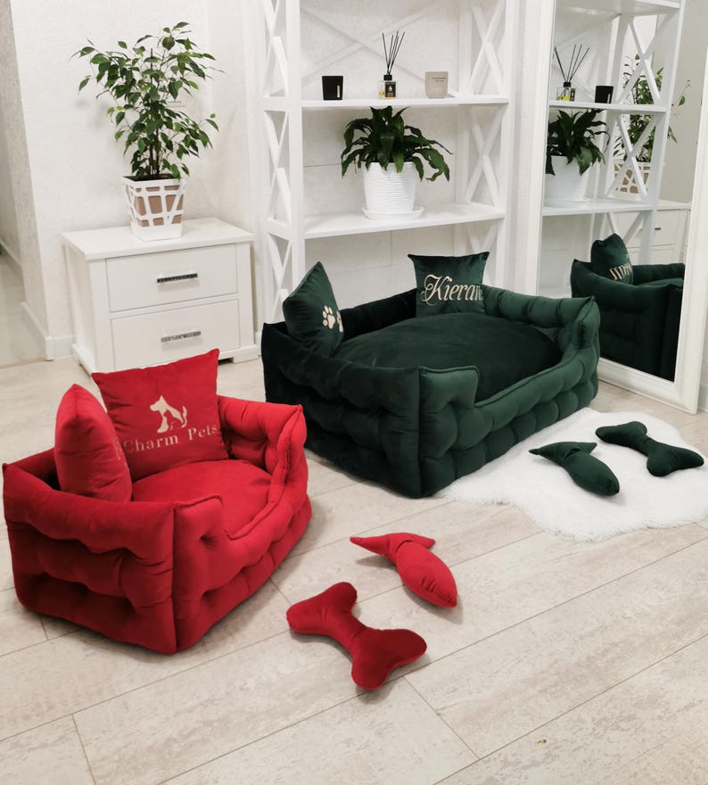 May include: Two pet beds, one red and one dark green, with matching pillows. The red bed has a pillow with the text "Charm Pets" and a dog silhouette. The green bed has a pillow with the name "Kieran". Several red and green bone-shaped toys are scattered around.