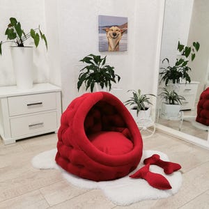 May include: Red velvet dog bed with a hooded design and a matching pillow. The bed is shaped like a dome and has a soft, plush interior.