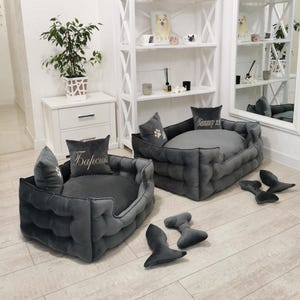 May include: Two gray, plush dog beds with matching pillows and toys. The beds have a tufted design and are rectangular. One pillow has a paw print, and the other has text. Several gray toys are scattered around the beds. The beds are on a light wood floor.
