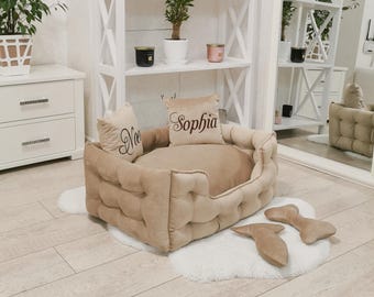 May include: A beige tufted dog bed with two matching bone-shaped pillows on a white faux fur rug. Two throw pillows with the names "Noe" and "Sophia" are on the bed. A white bookshelf and mirror are in the background.