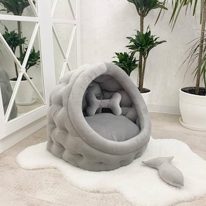 May include: A gray, tufted, round pet bed with a bone-shaped pillow inside. The bed is on a white fluffy rug.