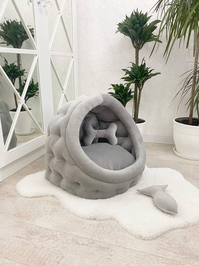 May include: A gray, tufted, round pet bed with a bone-shaped pillow inside. The bed is on a white fluffy rug.