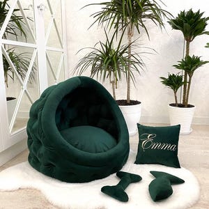 May include: A green velvet dog bed with a rounded, cave-like design. The bed has a plush, tufted surface and a removable cushion. The bed is accompanied by a matching green pillow with the name "Emma" embroidered on it and two green chew toys.