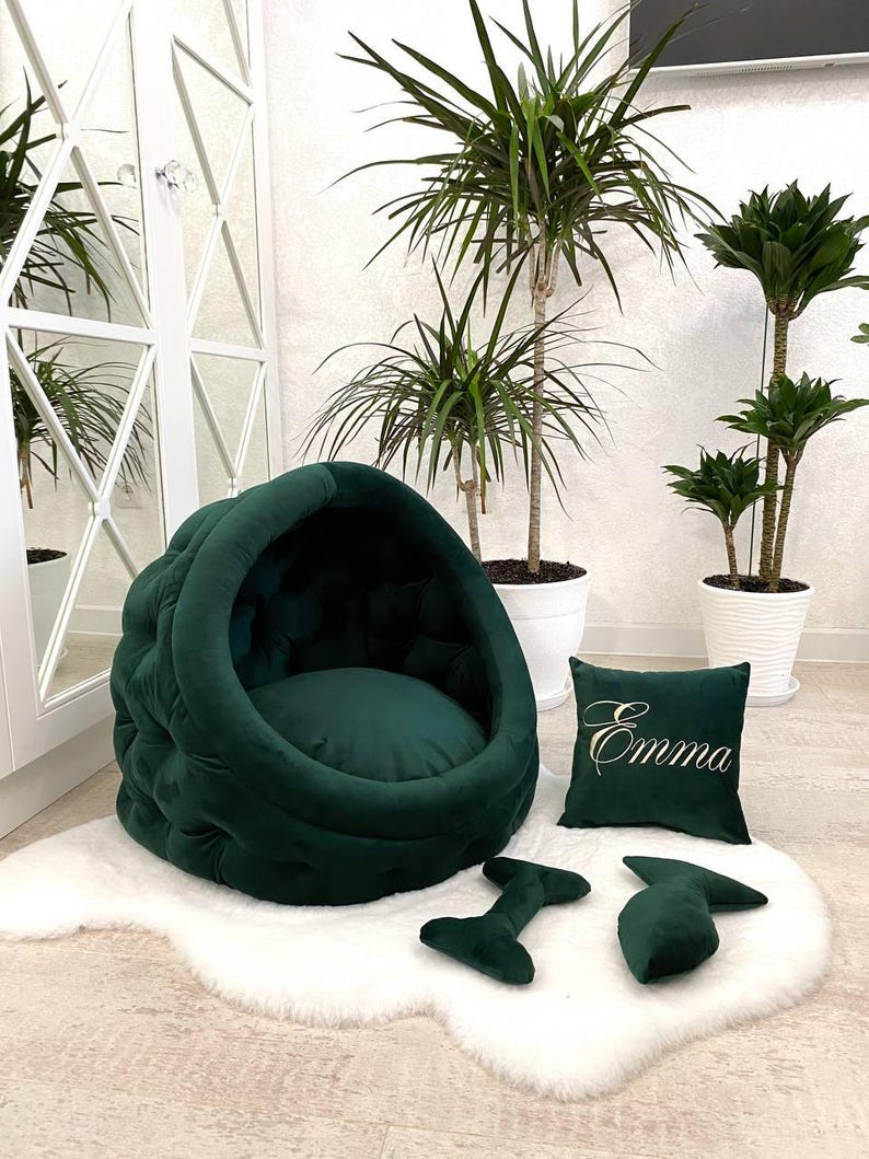 May include: A green velvet dog bed with a rounded, cave-like design. The bed has a plush, tufted surface and a removable cushion. The bed is accompanied by a matching green pillow with the name "Emma" embroidered on it and two green chew toys.