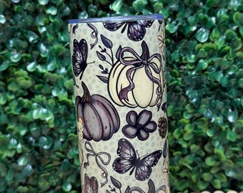 Puede incluir: Un vaso cilíndrico con un diseño de otoño. El vaso presenta ilustraciones de calabazas, mariposas y elementos florales en tonos morados, crema y marrón. El fondo es de color verde claro.