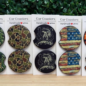 May include: Set of car coasters with various designs, including a deer in a forest, camouflage, a bear with a fish, an American flag, and a fish jumping out of water. Each coaster is round with a curved cutout. Text on the cards reads "Car Coasters Handmade with love."
