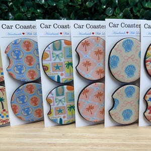 May include: A collection of car coasters with various colorful patterns, including palm trees, seashells, and lobsters. Each coaster has a black border and is displayed on a white card with the text "Car Coasters".