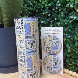 Può includere: Un bicchiere cilindrico e due sottobicchieri circolari per auto con un motivo ripetuto di anatre, fiocchi, lumache e zucche in blu, giallo e nero. La confezione dei sottobicchieri riporta il testo Car Coasters Handmade with love.