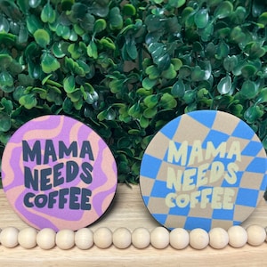 Könnte beinhalten: Zwei runde Getränkeuntersetzer. Einer hat ein lila-beiges Wellenmuster, der andere ein blau-beiges Karomuster. Beide Untersetzer tragen den Text "MAMA NEEDS COFFEE" in fetter Schrift.