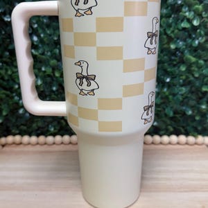 Puede incluir: Taza de viaje color crema con asa y tapa. La taza presenta un patrón repetido de gansos de dibujos animados con lazos, sobre un fondo a cuadros en color beige claro. El asa y la tapa son del mismo color crema.