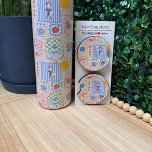 May include: A stainless steel tumbler and two car coasters with a whimsical pattern featuring geese, strawberries, and flowers. The tumbler is tall and cylindrical, while the coasters are round with a curved cutout. The text "Car Coasters Handmade with love" is printed on a white card.
