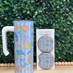 May include: A cylindrical tumbler with a white handle and a light blue striped pattern featuring colorful seashells, seahorses, jellyfish, and bows, alongside two circular car coasters on a card labeled Car Coasters Handmade with love.