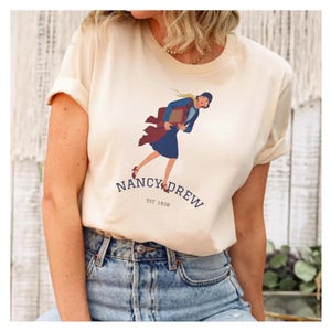 May include: A cream-colored short-sleeve t-shirt featuring a graphic illustration of a person in a blue dress and coat holding a book. Below the illustration, the text reads NANCY DREW and EST. 1930 in a dark blue font on the front of the shirt.