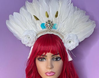 May include: A white feather headdress with a beaded crown, white fabric flowers, and gold accents. The headdress features a central jewel and turquoise accents. The mannequin has red hair and is set against a purple background.