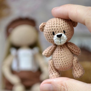 Op de afbeelding: Een kleine gehaakte teddybeer met een bruin lichaam en een wit gezicht. De beer wordt vastgehouden door een hand.