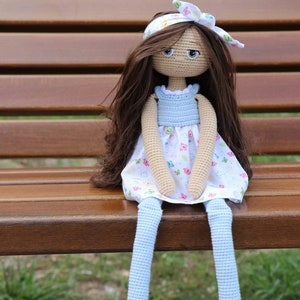 Puede incluir: Muñeca de ganchillo hecha a mano con pelo largo castaño, ojos azules y una diadema blanca con un lazo. La muñeca lleva un vestido azul claro con una falda blanca decorada con mariposas de colores y piernas azul claro.