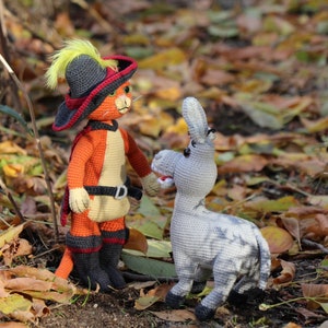 May include: A crocheted orange cat wearing a black and red hat with a yellow feather, standing next to a grey crocheted donkey in a pile of autumn leaves.