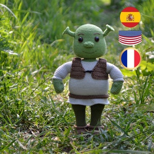 May include: A green crocheted plush toy of Shrek, a fictional ogre character, standing in a grassy area. The toy is wearing a brown vest and has a friendly expression. The background includes flags of Spain, the United States, and France.