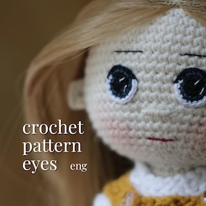Peut inclure: Patron de crochet pour les yeux de poupée, avec des instructions en anglais. L'image montre un gros plan sur le visage d'une poupée en crochet avec des yeux noirs et un léger fard à joues rose.