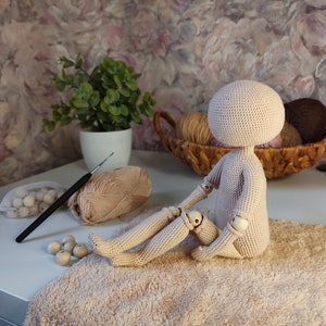 Peut inclure: Une poupée en crochet avec des articulations en bois, assise sur une serviette beige. La poupée est beige et a une tête ronde. La poupée est entourée de laine et d'autres fournitures d'artisanat.