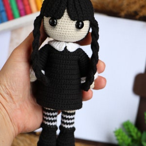 May include: Crocheted doll with black hair, a black dress, white collar, and black and white striped stockings. The doll is holding a book.
