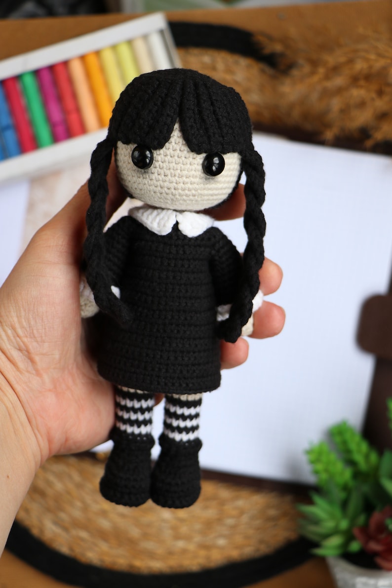 May include: Crocheted doll with black hair, a black dress, white collar, and black and white striped stockings. The doll is holding a book.
