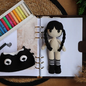 May include: A crocheted doll with black hair, a white body, and black and white striped socks. The doll is sitting on a notebook with a black and white crocheted hat beside it.