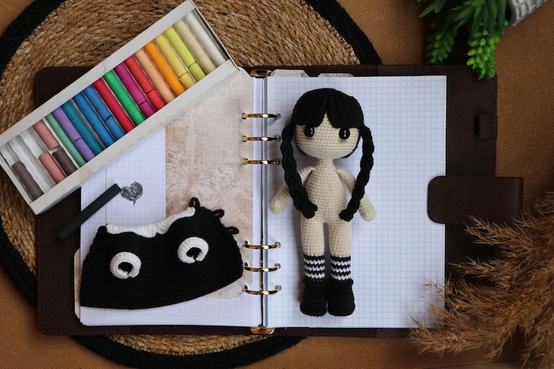 May include: A crocheted doll with black hair, a white body, and black and white striped socks. The doll is sitting on a notebook with a black and white crocheted hat beside it.