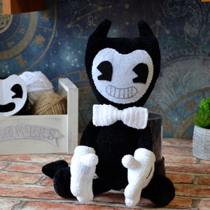 May include: Black and white crocheted plush toy of Bendy, a cartoon character with a bow tie, sitting on a brick surface.