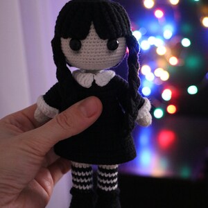 May include: A crocheted doll with black hair, a black dress, white collar, and white and black striped stockings. The doll is being held in a hand against a blurry background of colorful lights.