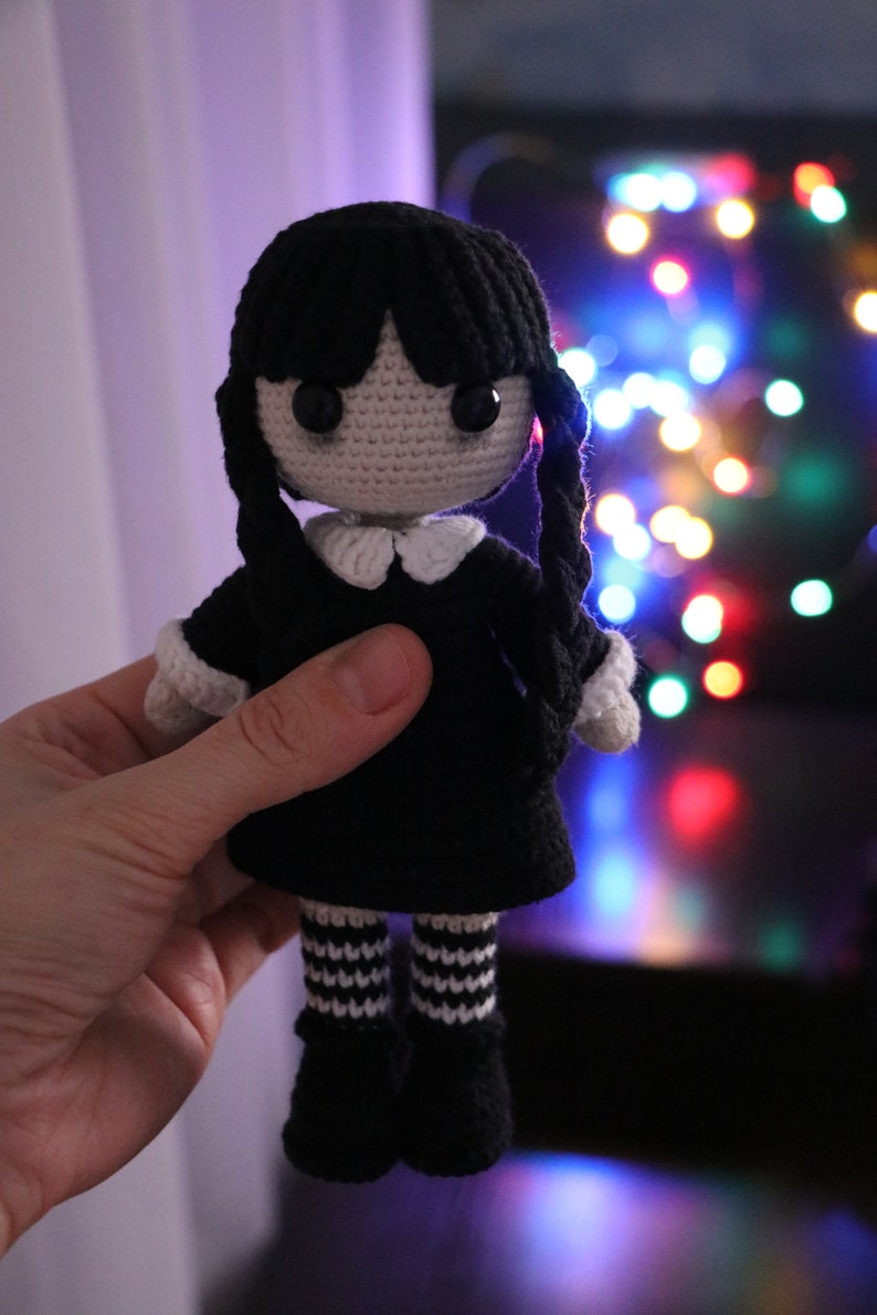 May include: A crocheted doll with black hair, a black dress, white collar, and white and black striped stockings. The doll is being held in a hand against a blurry background of colorful lights.