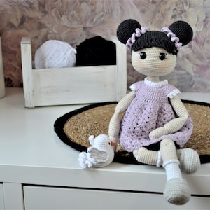 Peut inclure: Une poupée en crochet aux cheveux noirs, portant une robe violette et des chaussettes blanches, assise sur un tapis tissé avec un oiseau en crochet blanc.