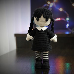 May include: A crocheted doll with black hair in pigtails, wearing a black dress with a white collar and black and white striped stockings. The doll has black eyes and a white face.