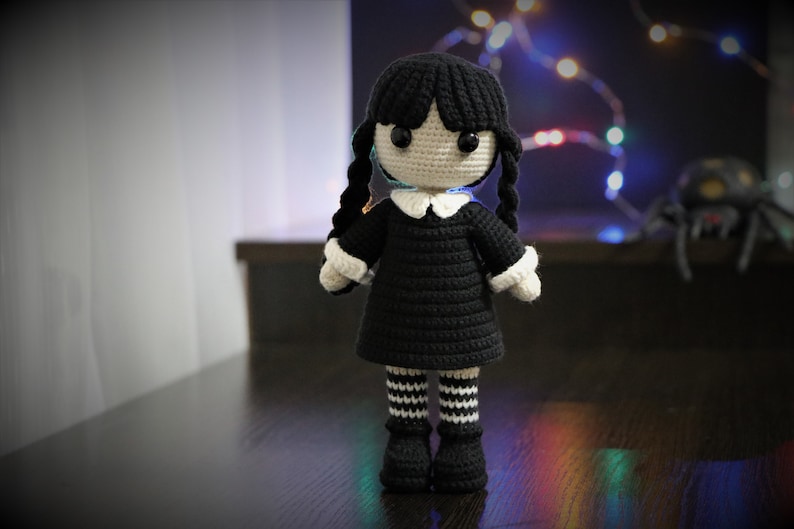 May include: A crocheted doll with black hair in pigtails, wearing a black dress with a white collar and black and white striped stockings. The doll has black eyes and a white face.