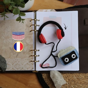 Könnte beinhalten: Ein gehäkeltes Kassettenband und Kopfhörer mit roten Ohrmuscheln liegen auf einem Notizbuch mit einem US-amerikanischen und französischen Flaggenaufkleber. Das Notizbuch ist geöffnet und hat einen goldenen Binder.