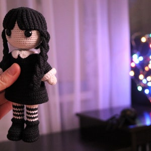 May include: A crocheted doll with black hair, a black dress, and white and black striped stockings. The doll is being held by a hand in front of a blurry background of a window and Christmas lights.