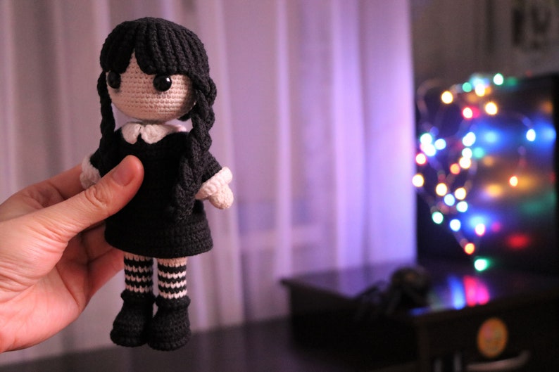 May include: A crocheted doll with black hair, a black dress, and white and black striped stockings. The doll is being held by a hand in front of a blurry background of a window and Christmas lights.