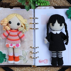 May include: Two crocheted dolls, one with blonde hair and a pink and orange striped sweater, the other with black hair and a black dress, are displayed on a page of a notebook.