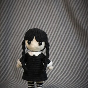 May include: A crocheted doll with black hair, a black dress, white collar, and black and white striped stockings. The doll is standing on a gray striped surface.