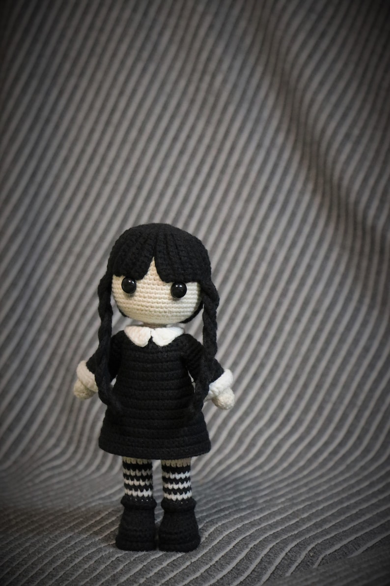 May include: A crocheted doll with black hair, a black dress, white collar, and black and white striped stockings. The doll is standing on a gray striped surface.