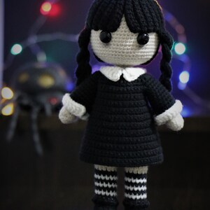 May include: A crocheted doll with black hair, a black dress, white collar, and black and white striped stockings. The doll is standing on a dark surface.