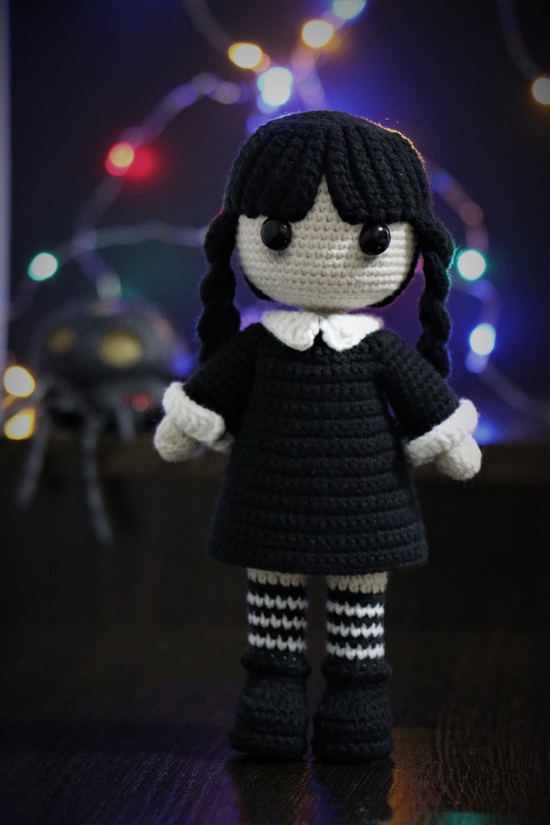 May include: A crocheted doll with black hair, a black dress, white collar, and black and white striped stockings. The doll is standing on a dark surface.